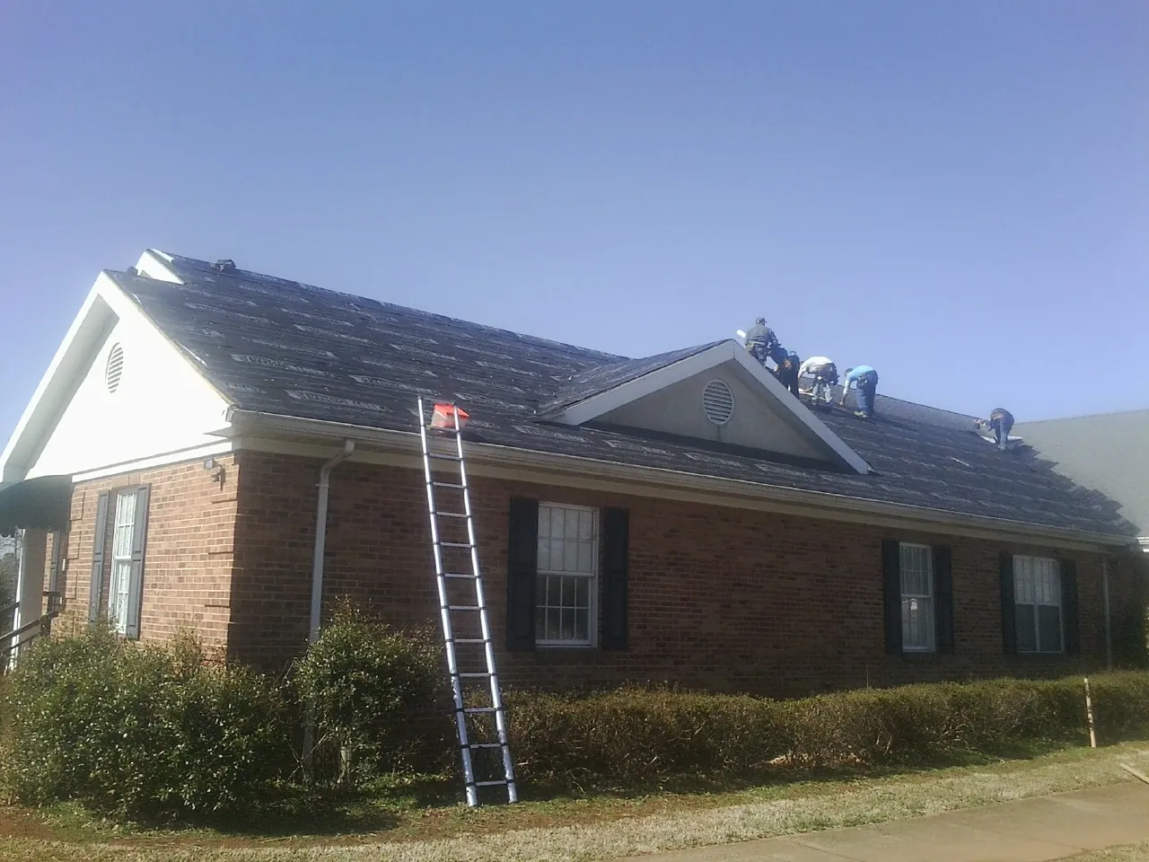 Crew tearing shingles off a brick ranch roof with a ladder set — Emergency Roof Repair work by our crew in Lutz